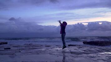 Girl on the seashore against the backdrop of storm waves. She is happy and enjoys the fresh wind. 4K video