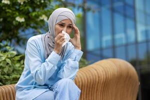 Sad muslim female sitting on wooden bench and covering face with paper tissue for blowing out. Sick woman experiencing allergy to pollen and trying hiding unpleasant symptoms of running nose. photo