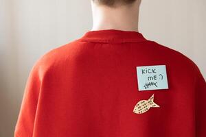 Boy in red hoodie with a paper attached with tape to his back with the handwriting text kick me. Fools day joke photo