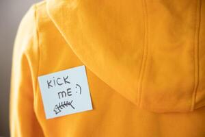 Boy in yellow hoodie with a sticky note on his back, saying kick me. photo