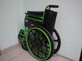 a green folding wheelchair leaning against a white wall. The wheelchair is titled to the side with the left wheel resting against the wall photo