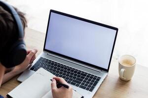 man in wireless headphones makes notes in front of laptop with blank screen, notebook and coffee nearby photo