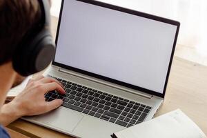 man in wireless headphones types on a laptop, notebook nearby, blank screen photo