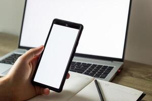 man holds mobile phone at the table in front of laptop, blank screens photo