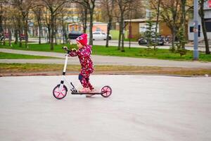 alegre activo niño paseos un rosado scooter en un ciudad parque foto