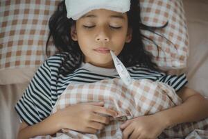 asiático madre medición temperatura niña con digital termómetro en su boca en cama a Mañana tiempo, enfermo niño tener frio toalla para reducir alto fiebre, foto