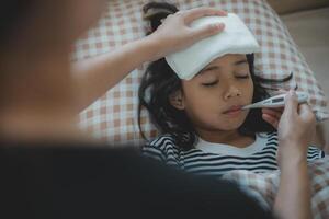 asiático madre medición temperatura niña con digital termómetro en su boca en cama a Mañana tiempo, enfermo niño tener frio toalla para reducir alto fiebre, foto