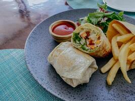 Tortilla wrap with fried chicken meat and vegetables served with sauce and french fries on a wooden table. photo