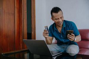 Happy young businessman smiling holding credit card while reading his smartphone. Smiling businessman reading messages with his cell phone. Man working at his desk in the office. photo