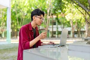 Young Asian man smiling while using laptop for working and conference meeting at the morning. Asian man having a call on laptop. Concept of remote and freelance photo