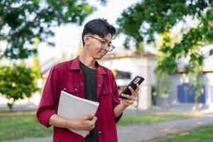 Young Asian man using smartphone with happy expression. A male smiling while holding his phone at the public park photo