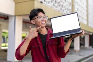 Young Asian man wearing red shirt smiling and showing white laptop screen for advertisement. Concept of people lifestyle. Happy man holding blank laptop screen with blurred background. photo