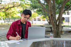 Asian college student studying on laptop at campus outdoor park. Man writing on a note book and working on laptop. Educational concept photo