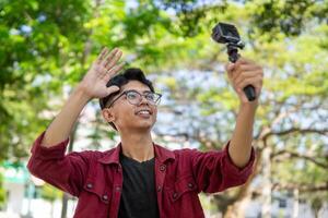 asiático hombre grabación y hablando a cámara. joven hombre vlogging y tomando selfie con acción cámara a público parque. foto