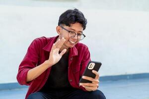 Asian man holding handphone and calling with friends waving hand making hello gesture. Man using smartphone for online conference call photo