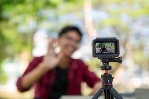 asiático hombre grabación y hablando a cámara. joven hombre vlogging y tomando selfie con acción cámara a público parque. acción cámara grabación un hombre con borroso antecedentes foto