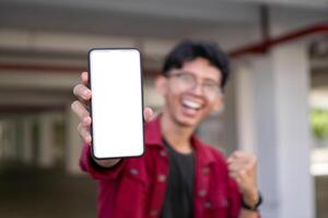 joven asiático hombre vistiendo rojo camisa sonriente y demostración blanco móvil teléfono pantalla para anuncio publicitario. concepto de personas estilo de vida. contento hombre participación blanco teléfono inteligente pantalla con borroso antecedentes. foto