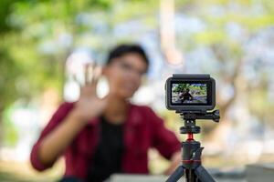 asiático hombre grabación y hablando a cámara. joven hombre vlogging y tomando selfie con acción cámara a público parque. acción cámara grabación un hombre con borroso antecedentes foto