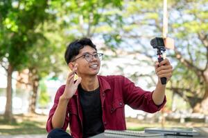 asiático hombre grabación y hablando a cámara. joven hombre vlogging y tomando selfie con acción cámara a público parque. foto