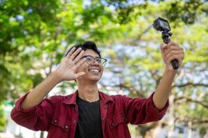 Asian man recording and talking to camera. Young man vlogging and taking selfie with action camera at public park. photo