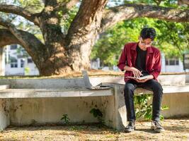 asiático Universidad estudiante estudiando en ordenador portátil a instalaciones al aire libre parque. hombre escritura en un Nota libro y trabajando en ordenador portátil. educativo concepto foto