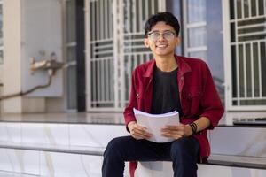 Asian college student smiling and looking. Portrait of a college student at campus. Asian male student sitting while holding a book looking at the camera. photo