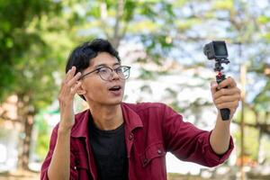 Asian man recording and talking to camera. Young man vlogging and taking selfie with action camera at public park. photo