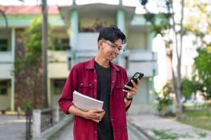 Young Asian college student using smartphone with happy expression. A male smiling while holding his phone and books at the public park. copy space photo