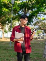 Young Asian college student using smartphone with happy expression. A male smiling while holding his phone and books at the public park. copy space photo