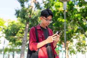 joven asiático Universidad estudiante utilizando teléfono inteligente con contento expresión. un masculino sonriente mientras participación su teléfono y libros a el público parque. Copiar espacio foto