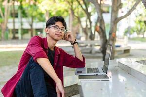 joven asiático hombre sentado al aire libre con contento expresión. un masculino Universidad estudiante sonriente a el público parque foto