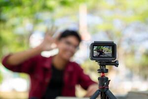 asiático hombre grabación y hablando a cámara. joven hombre vlogging y tomando selfie con acción cámara a público parque. acción cámara grabación un hombre con borroso antecedentes foto