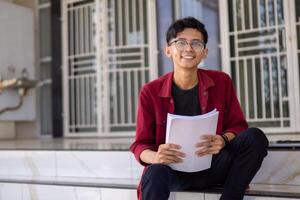 Asian college student smiling and looking. Portrait of a college student at campus. Asian male student sitting while holding a book looking at the camera. photo