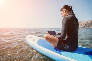 Woman sea laptop. Digital nomad, freelancer with laptop working on sup board at calm sea beach. Happy smiling girl relieves stress from work. Freelance, digital nomad, travel and holidays concept photo