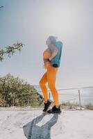 Fitness woman sea. Happy middle aged woman in orange sportswear exercises morning outdoors on yoga mat with laptop in park over ocean beach. Female fitness pilates yoga routine. Healthy lifestyle. photo