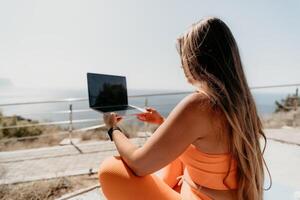 Woman laptop sea. Working remotely on seashore. Happy successful woman female freelancer working on laptop by the sea at sunset, makes a business transaction online. Freelance, remote work on vacation photo