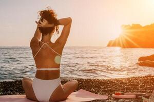 Woman sea yoga. Happy woman in white swimsuit and boho style braclets practicing outdoors on yoga mat by sea on sunset. Women yoga fitness routine. Healthy lifestyle, harmony and meditation photo