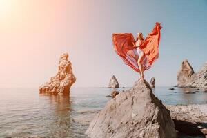 Woman travel sea. Young Happy woman in a long red dress posing on a beach near the sea on background of volcanic rocks, like in Iceland, sharing travel adventure journey photo