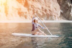Woman sup yoga. Middle age sporty woman practising yoga pilates on paddle sup surfboard. Female stretching doing workout on sea water. Modern individual female hipster outdoor summer sport activity. photo