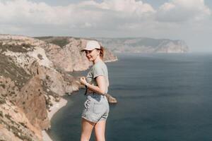 mujer viaje mar. contento turista en sombrero disfrutar tomando imagen al aire libre para recuerdos. mujer viajero posando en el playa a mar rodeado por volcánico montañas, compartiendo viaje aventuras viaje foto