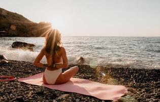 Woman sea yoga. Happy woman in white swimsuit and boho style braclets practicing outdoors on yoga mat by sea on sunset. Women yoga fitness routine. Healthy lifestyle, harmony and meditation photo