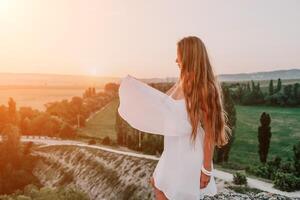 Happy woman standing with her back on the sunset in nature in summer with open hands. Romantic beautiful bride in white boho dress posing with mountains on sunset photo