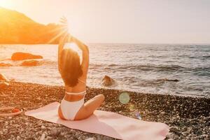 Woman sea yoga. Happy woman in white swimsuit and boho style braclets practicing outdoors on yoga mat by sea on sunset. Women yoga fitness routine. Healthy lifestyle, harmony and meditation photo