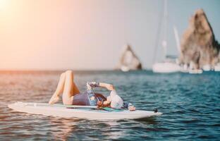 Woman sup yoga. Middle age sporty woman practising yoga pilates on paddle sup surfboard. Female stretching doing workout on sea water. Modern individual female hipster outdoor summer sport activity. photo