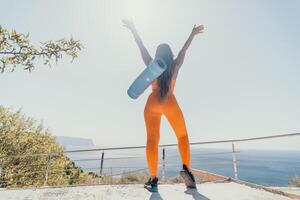 Fitness woman sea. Happy middle aged woman in orange sportswear exercises morning outdoors on yoga mat with laptop in park over ocean beach. Female fitness pilates yoga routine. Healthy lifestyle. photo