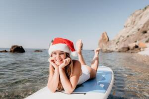 mujer mar sorber. cerca arriba retrato de contento joven caucásico mujer con largo pelo en Papa Noel sombrero mirando a cámara y sonriente. linda mujer retrato en un blanco bikini posando en cenar tablero en el mar foto