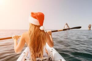 Woman in kayak back view. Happy young woman in Santa hat floating in kayak on calm sea. Summer holiday vacation and cheerful female people relaxing having fun on the boat. photo