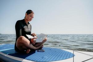 Woman sea laptop. Digital nomad, freelancer with laptop working on sup board at calm sea beach. Happy smiling girl relieves stress from work. Freelance, digital nomad, travel and holidays concept photo