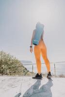 Fitness woman sea. Happy middle aged woman in orange sportswear exercises morning outdoors on yoga mat with laptop in park over ocean beach. Female fitness pilates yoga routine. Healthy lifestyle. photo