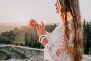 Happy woman in white boho dress on sunset in mountains. Romantic woman with long hair standing with her back on the sunset in nature in summer with open hands. Silhouette. Nature. Sunset. photo